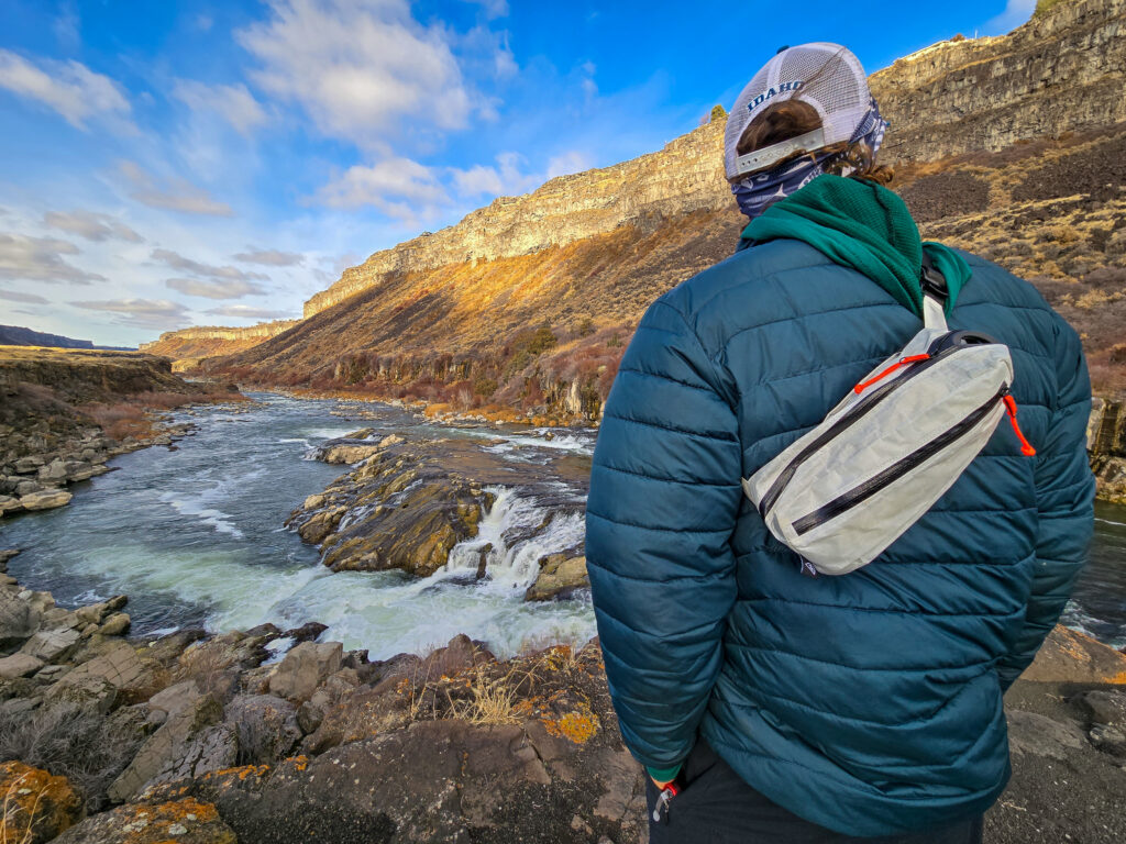 a man wearing a fanny pack as a shoulder sling next to a rushing river 