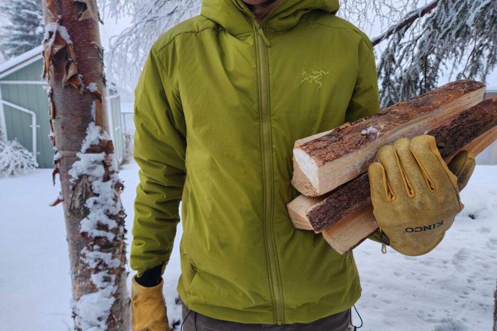A person in a green jacket and yellow gloves stands in a snowy yard holding firewood, surrounded by snow-covered trees.