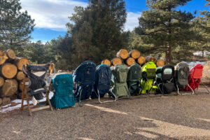 10 baby backpack carriers lineup up in front of a wood pile with pine trees in the background.