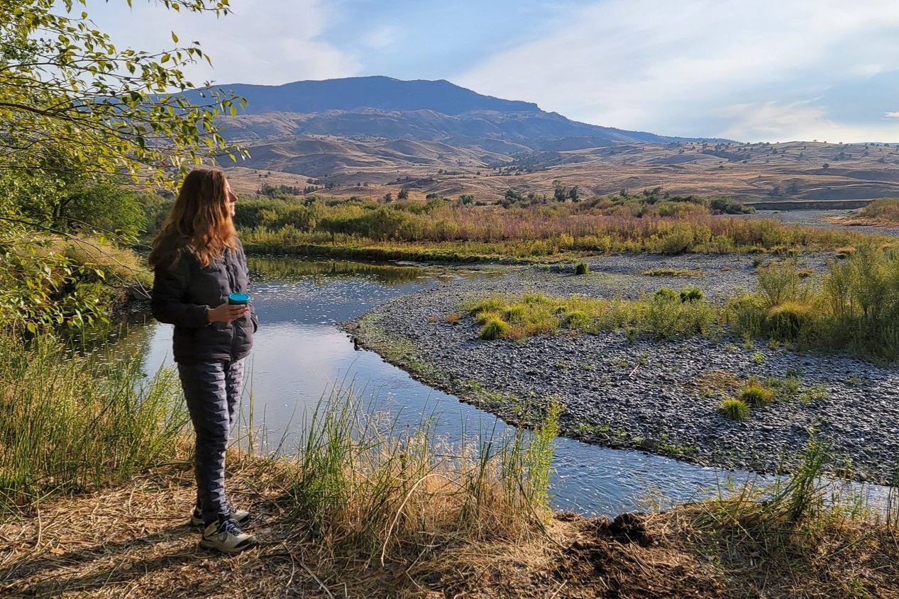 a woman drinking coffee while wearing a warm puffy jacket while looking at a river and mountain scene
