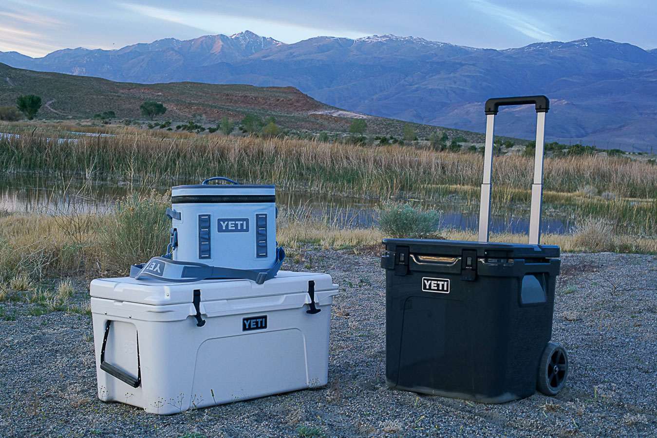 The YETI Hopper Flip 12 soft shell cooler sits on top of the YETI Tundra 65 cooler. The YETI Roadie 48 cooler sits to the right with mountains and a body of water in the background.