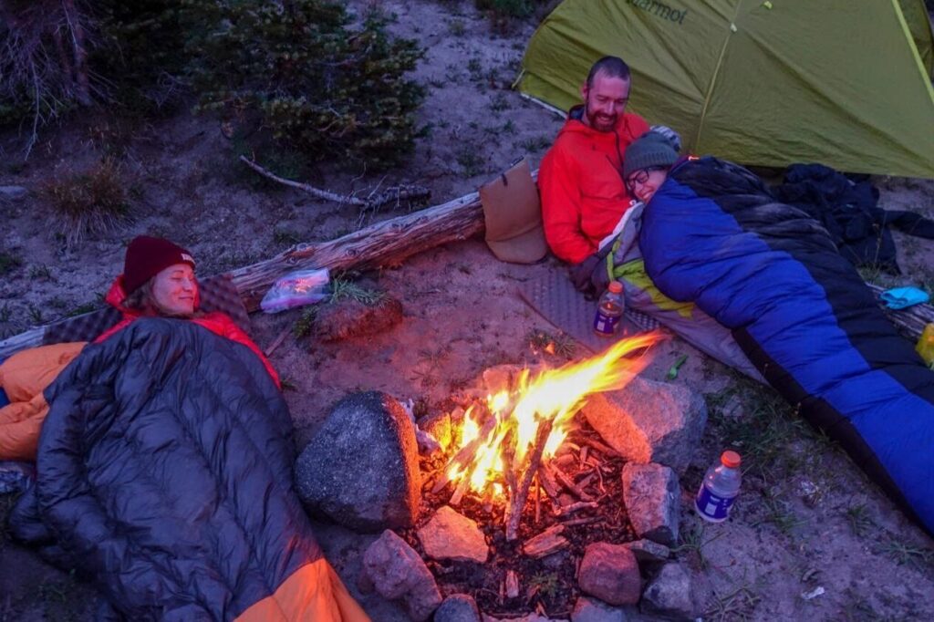 campers in their sleeping bags next to a fire