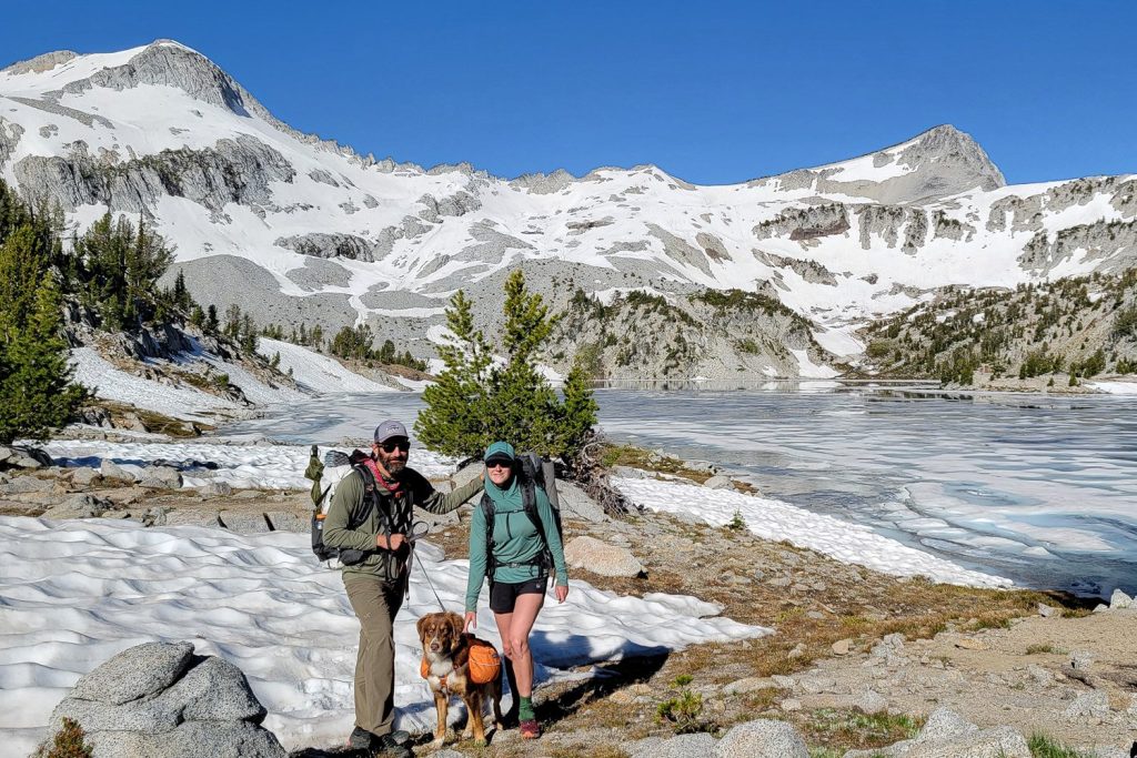 two hikers and their dog posing in front of a frozen alpine lake on a cloudless day