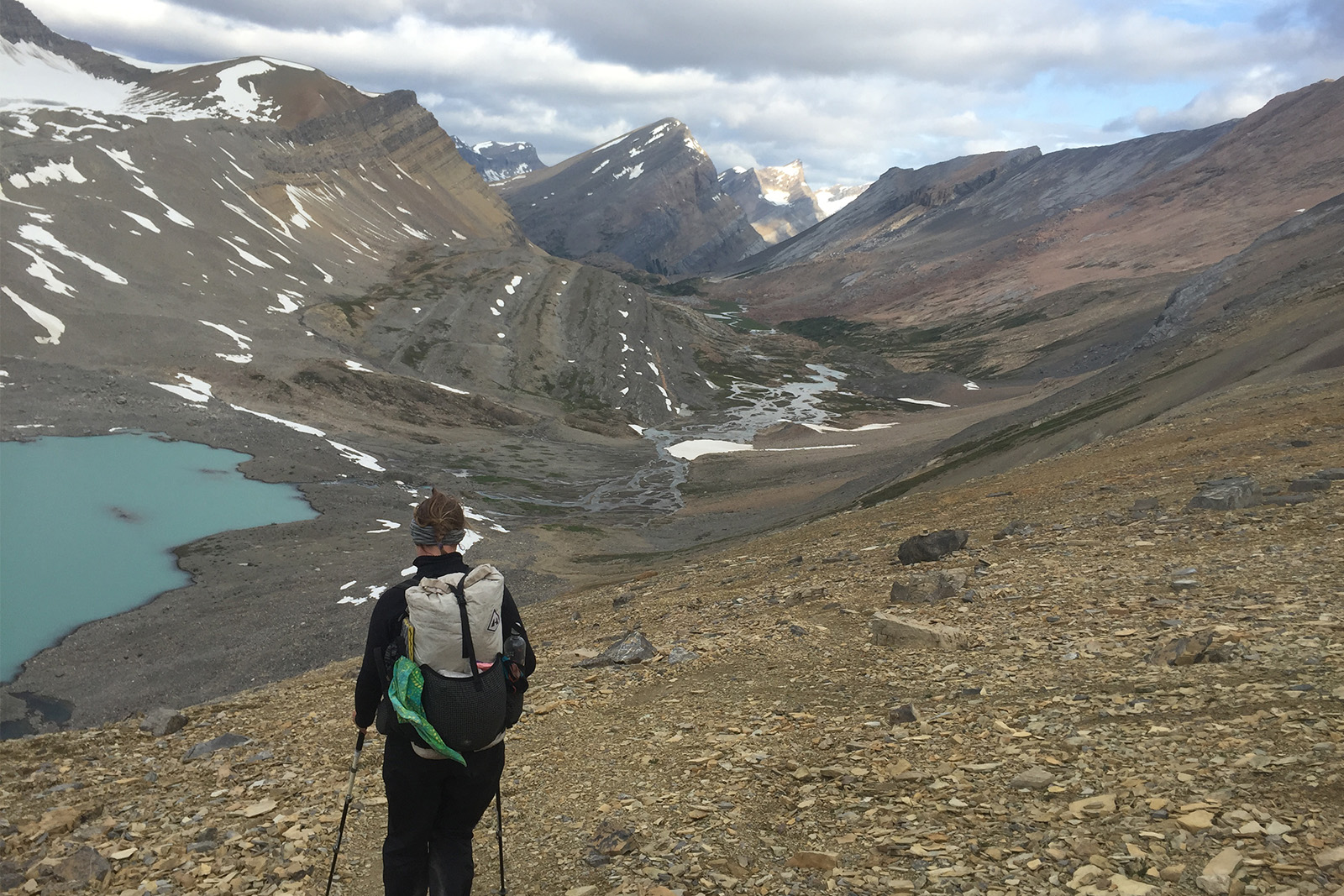 A person wearing a pack is hiking down a scree slope. There is a glacial blue lake in the valley below and mountains in the distance.