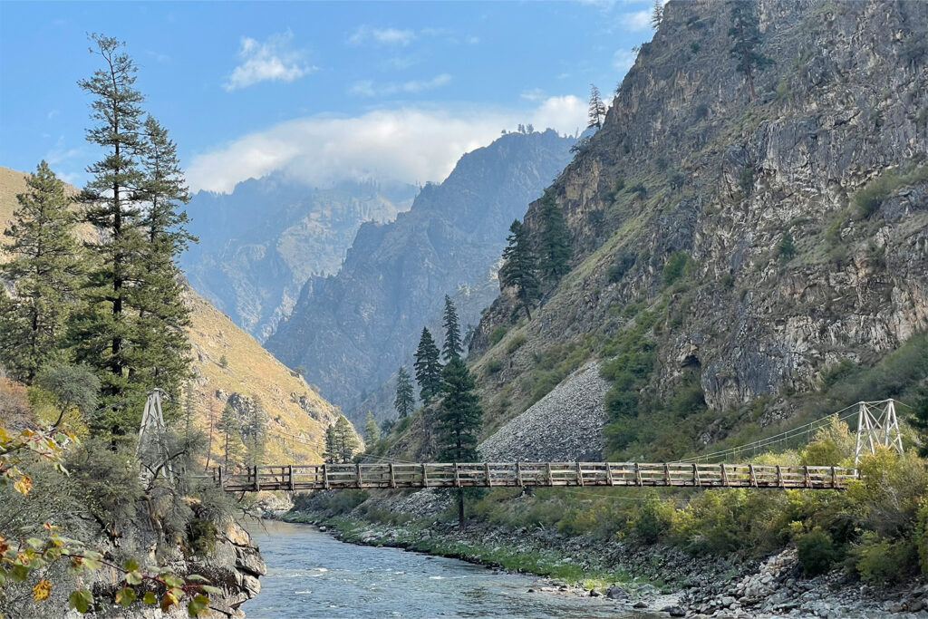 Landscape photo of a wooden suspension bridge spanning a good sized river. Beyond the bridge, granite peaks rise steeply making the valley deep cut.