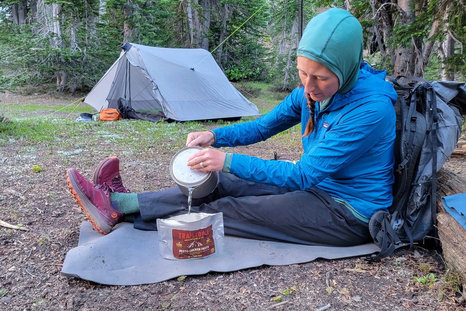 backpacker sitting on a foam pad, resting their back on an emptied backpack against a log. They are pouring hot water into a dehydrated meal pouch with their tent set up in the background.
