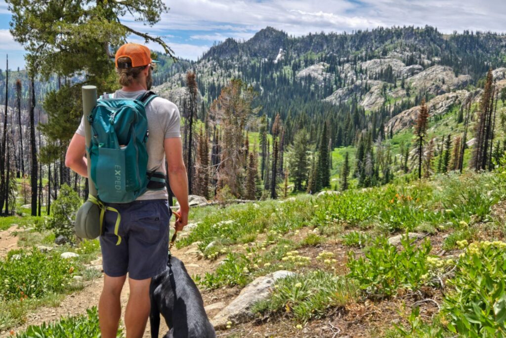 A person with a black dog looks out over a rocky landscape filled with pines. An EXPED brand pack is on their bag and a fly fishing rod is strapped to the side.