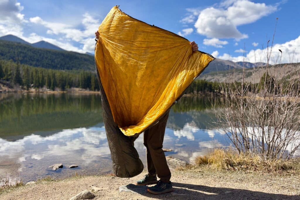 a hiker holds up a grey quilt to show the yellow inside to the camera. a lake a mountains are in the background.