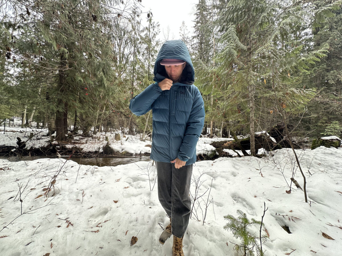 women in a snowy forest near a small creek zipping up a winter down jacket