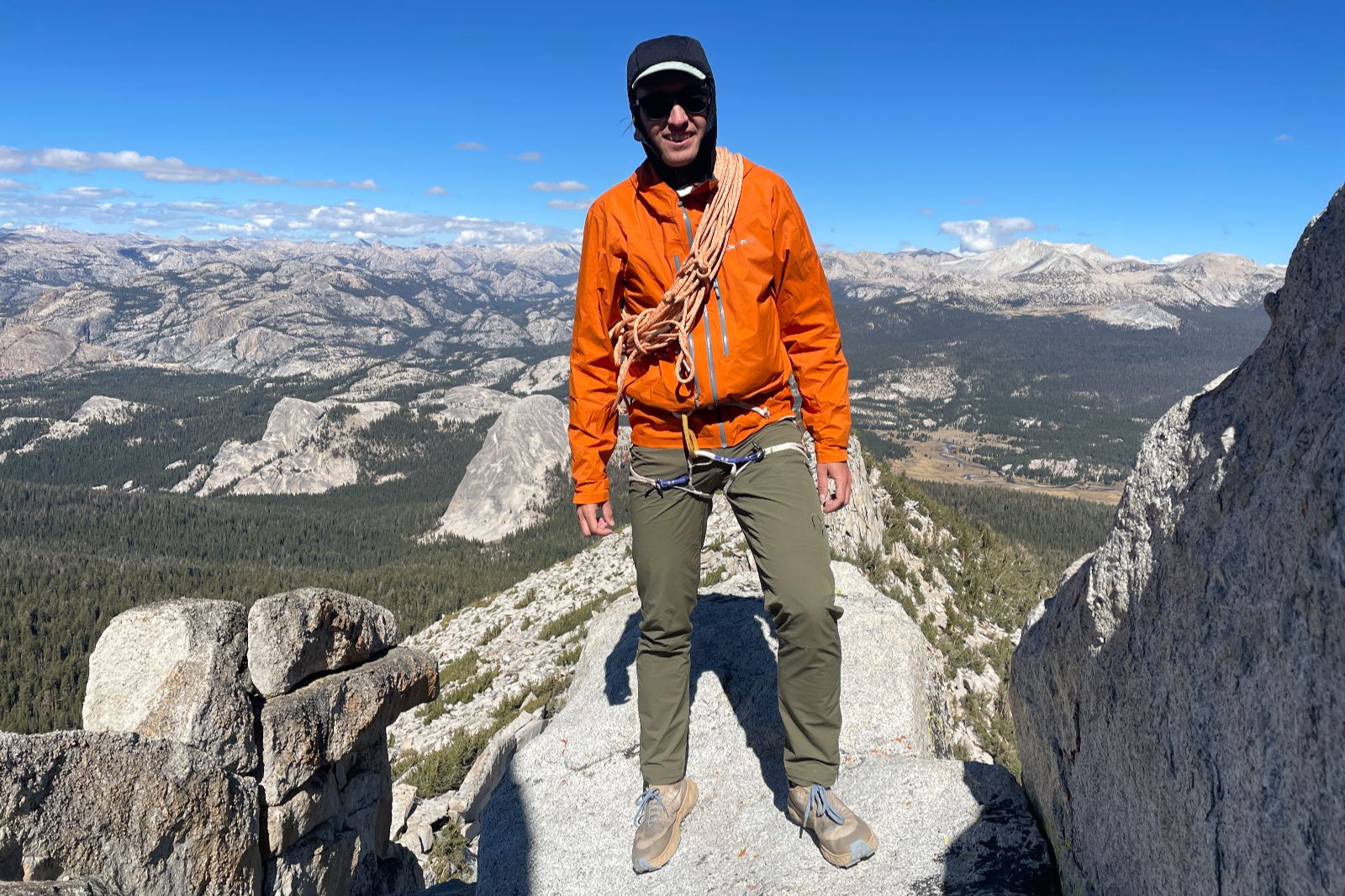 A man standing on an alpine ridge wearing an orange jacket with a rope couled around his shoulders.