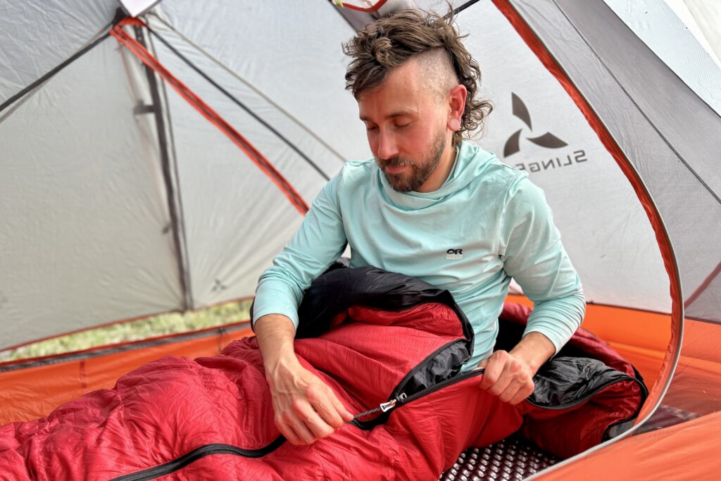 A man messes with the zipper on a red sleeping bag inside of a tent.
