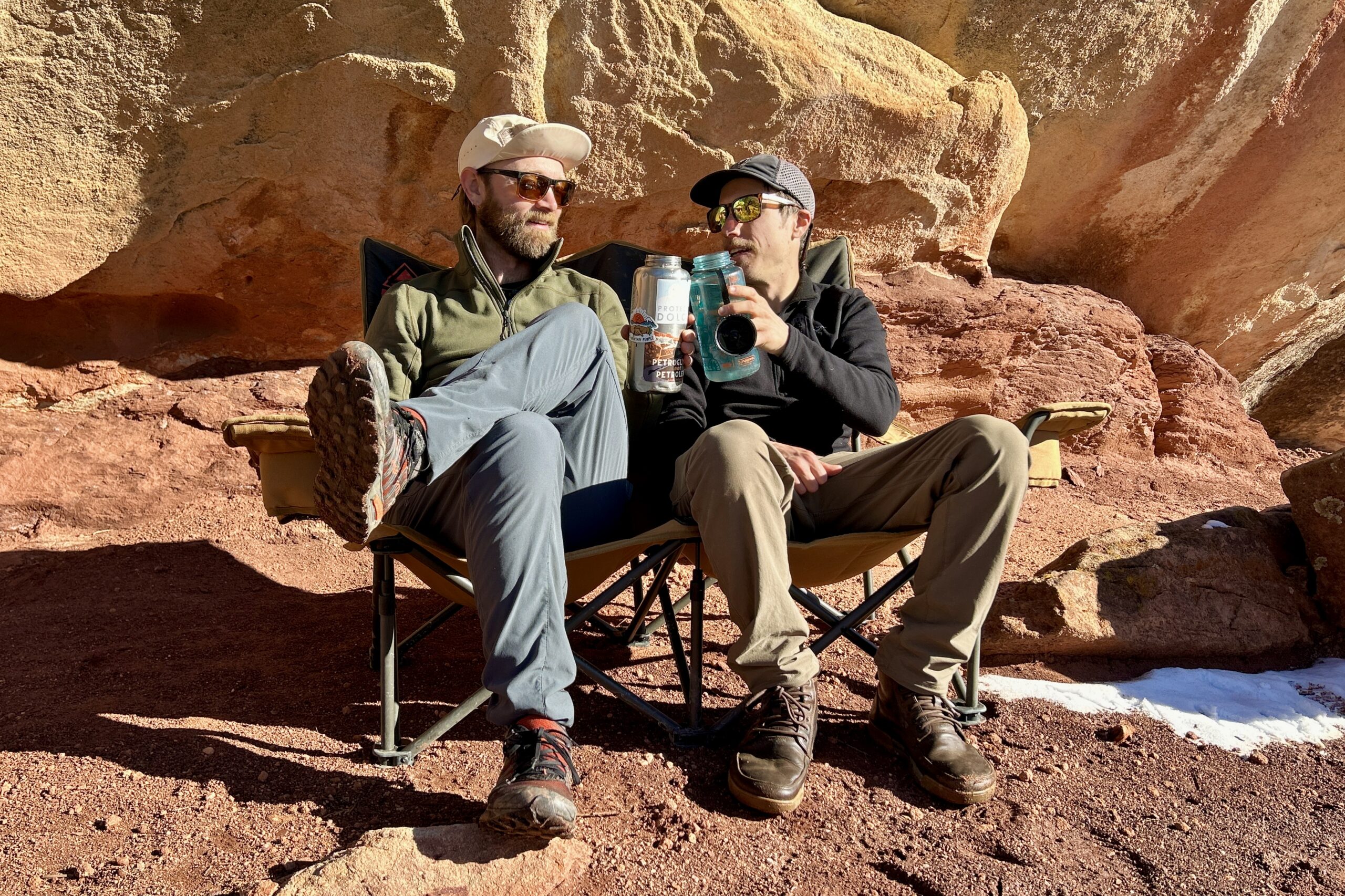 Two men cheers their water bottles while sitting next to each other on a 2-person camping chair.