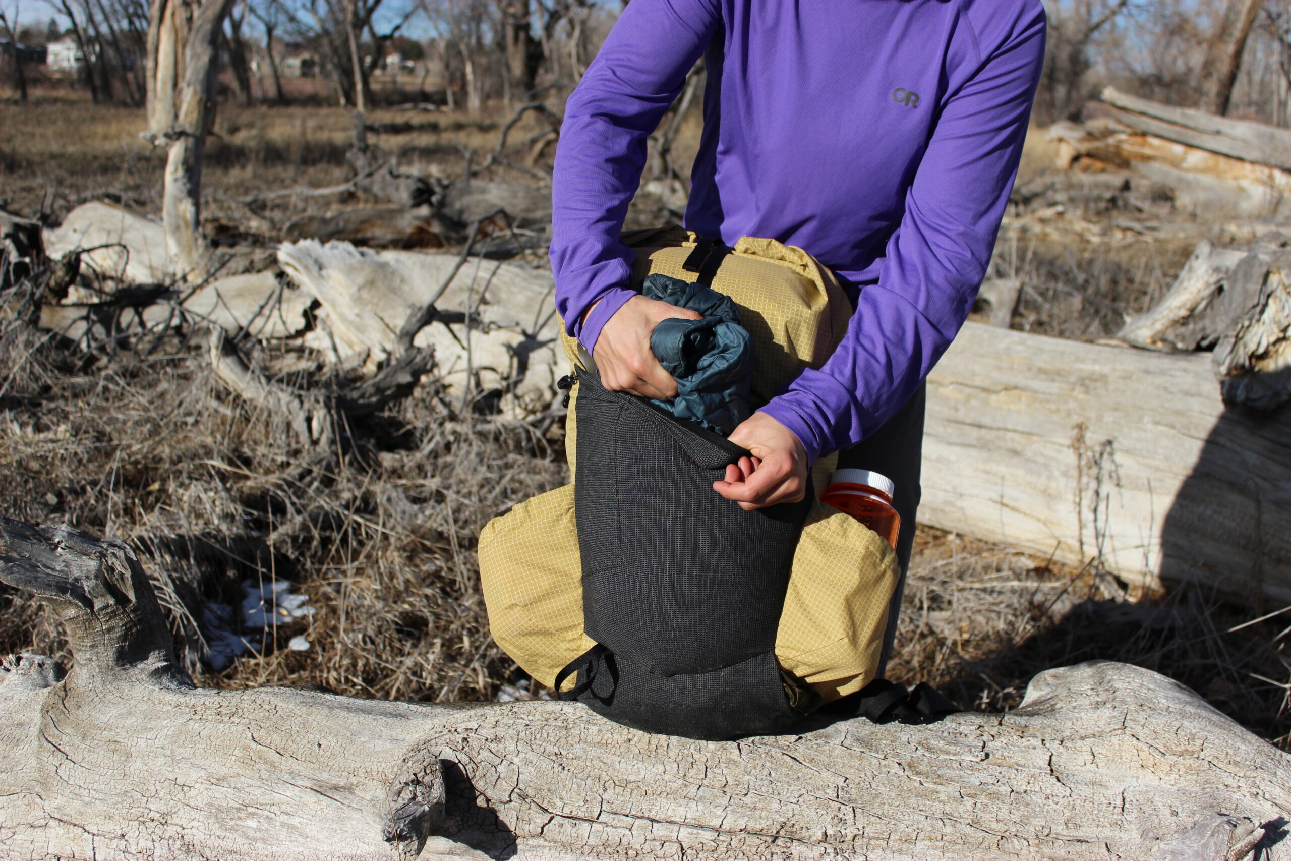 A hiker pulls a jacket out of the front pocket of a yellow backpack sitting on a log.