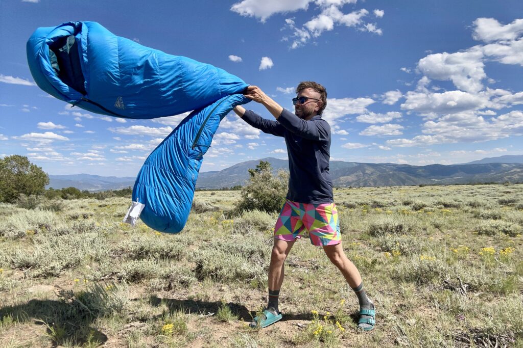 A man shakes out a long sleeping bag in a field with mountains and blue sky in the background.