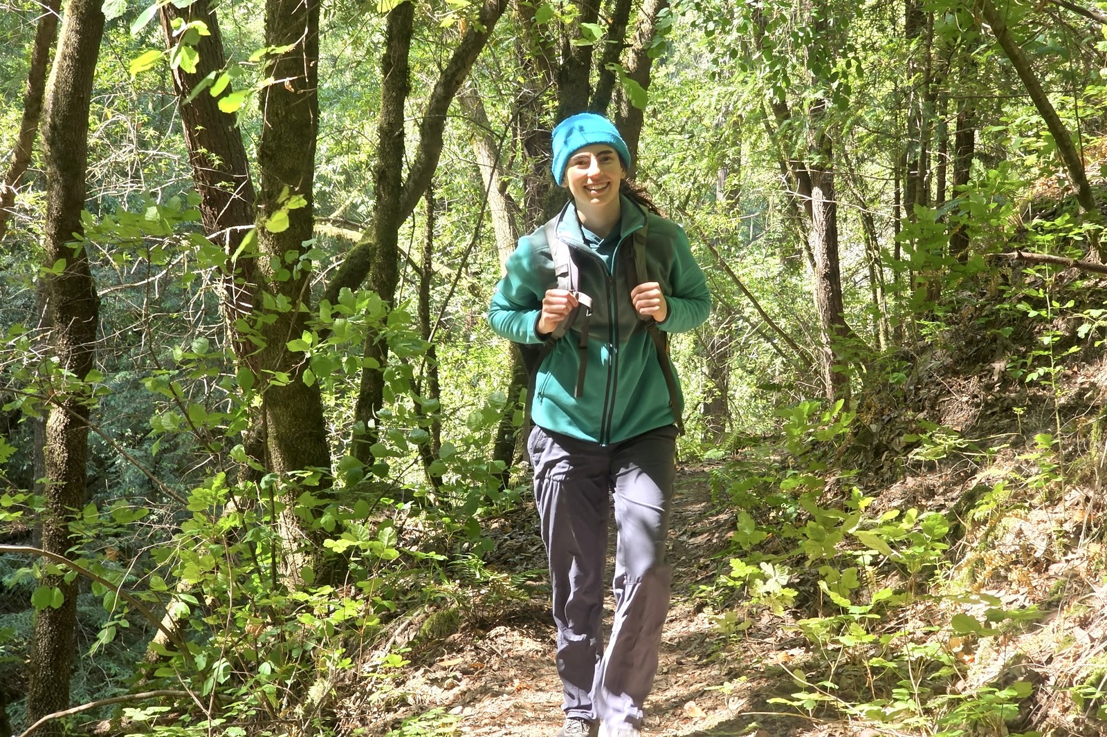 A woman hikes through a sun-dappled forest wearing a pack over her fleece jacket.