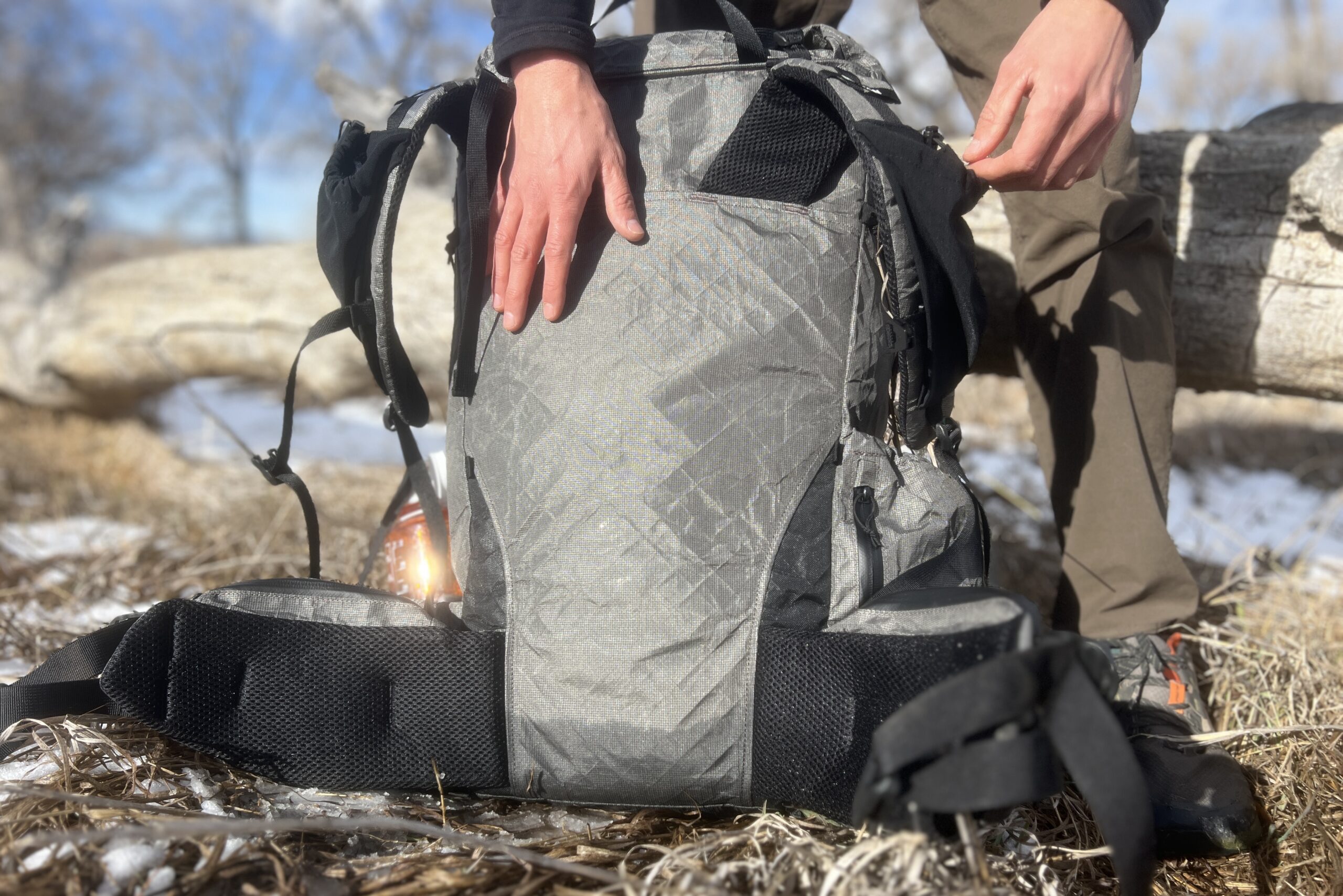 A hiker puts their hand on the back panel of a grey backpack with a log in the background.