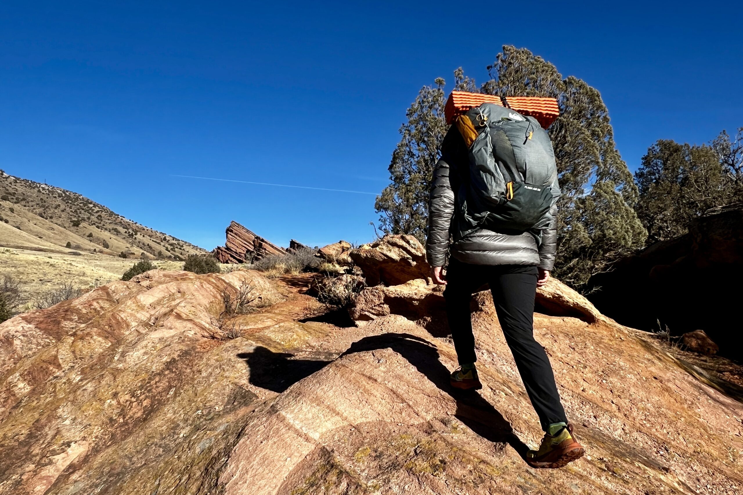 A hiker walks up a red rock formation while wearing a grey backpack.