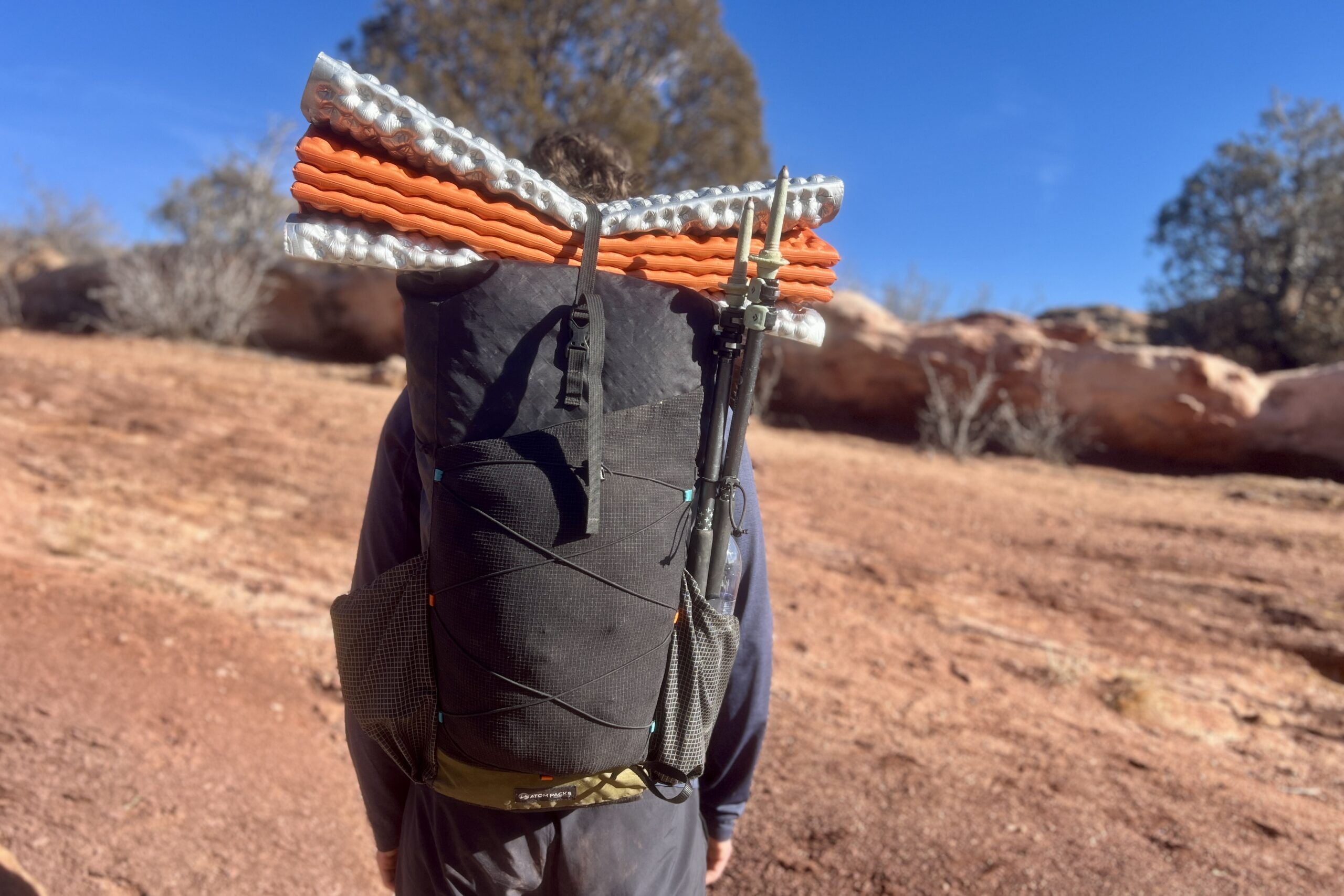 Back view of the Atom Packs backpack with orange and white sleeping pad secured on top and trekking poles mounted to the side.