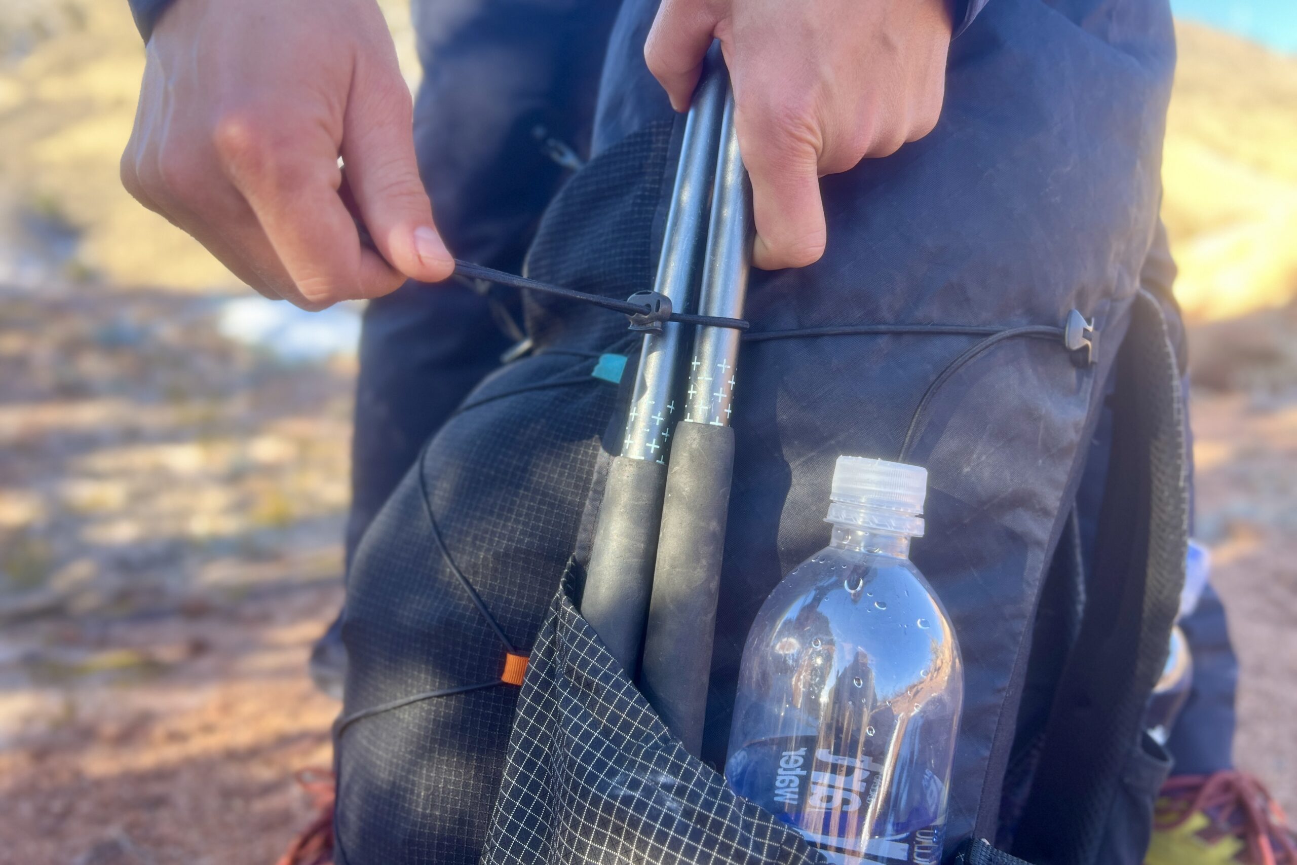 Close-up detail of a backpack's side pocket showing how trekking poles and a water bottle can be secured simultaneously using built-in compression straps against a black gridstop material.