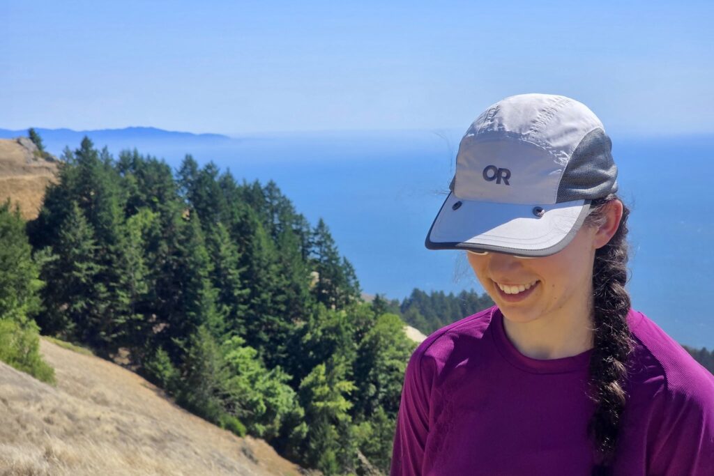 A woman wears a hat as she hikes on a grassy hill over the ocean.