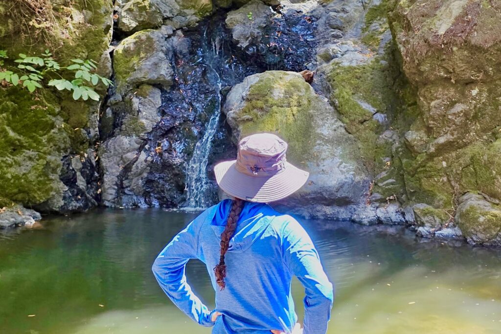 A woman stands before a waterfall wearing her sun hat.