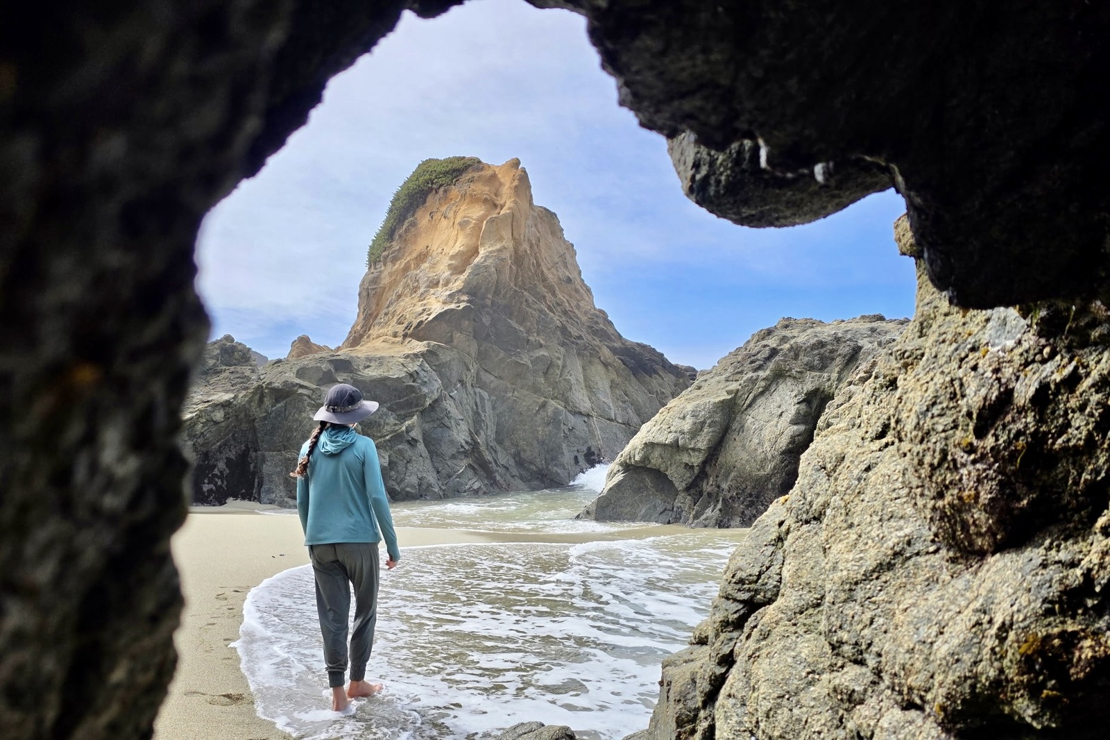A woman walks along a beach with large rock formations in the background.