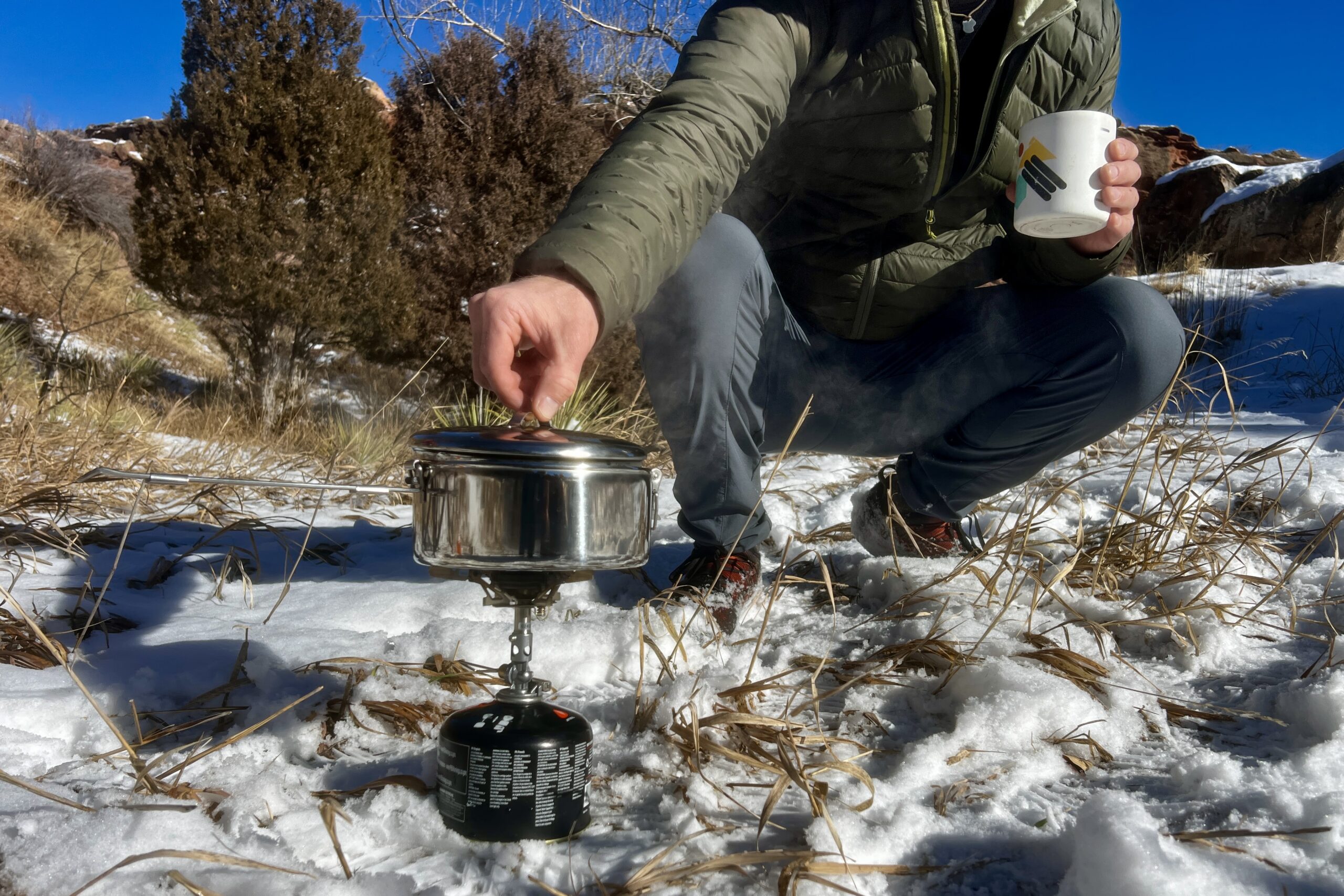 A hiker crouches next to a backpacking pot cooking on a stove and begins to lift the lid.