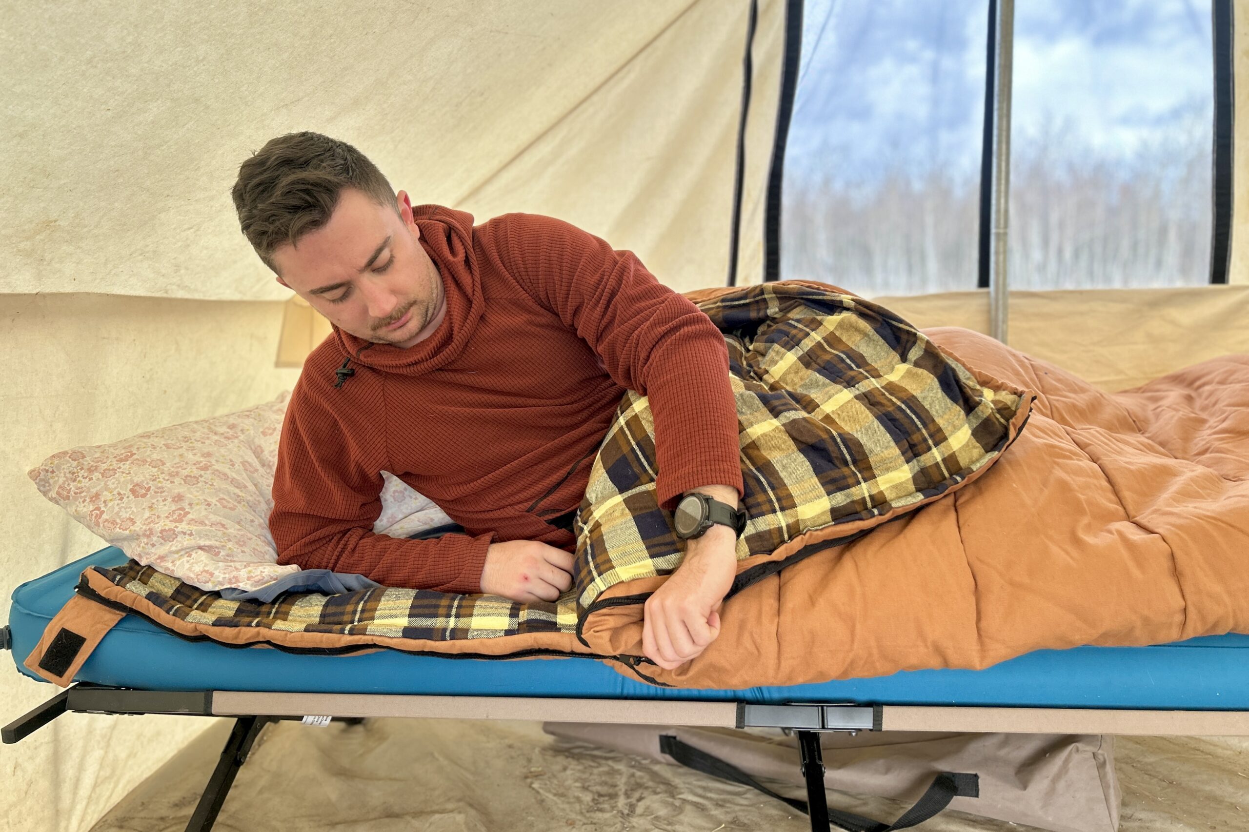 A man leans on one arm while laying on a sleeping bag, unzipping the side.