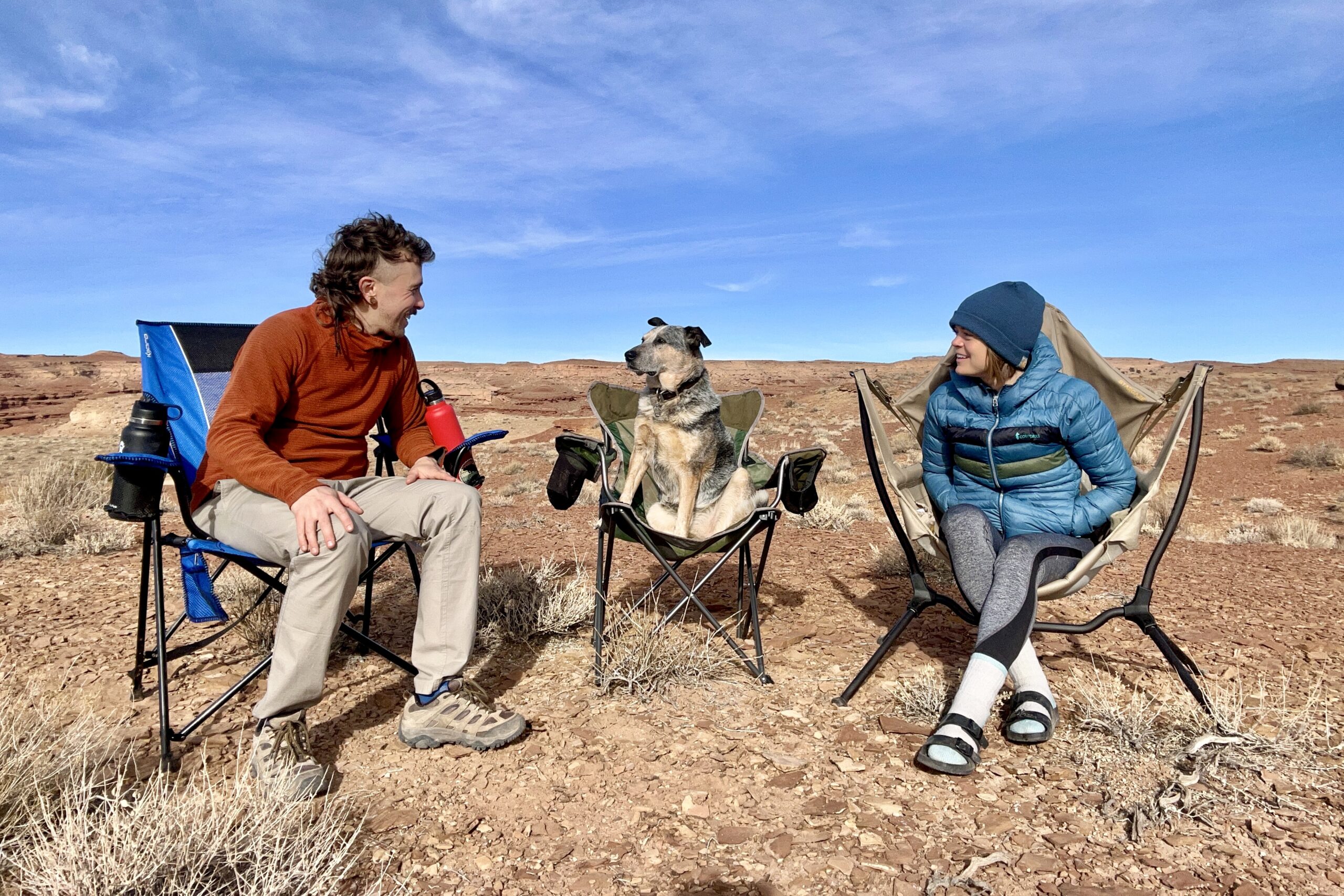 A man, woman, and dog each sit in their own camping chairs in a sunny desert setting.
