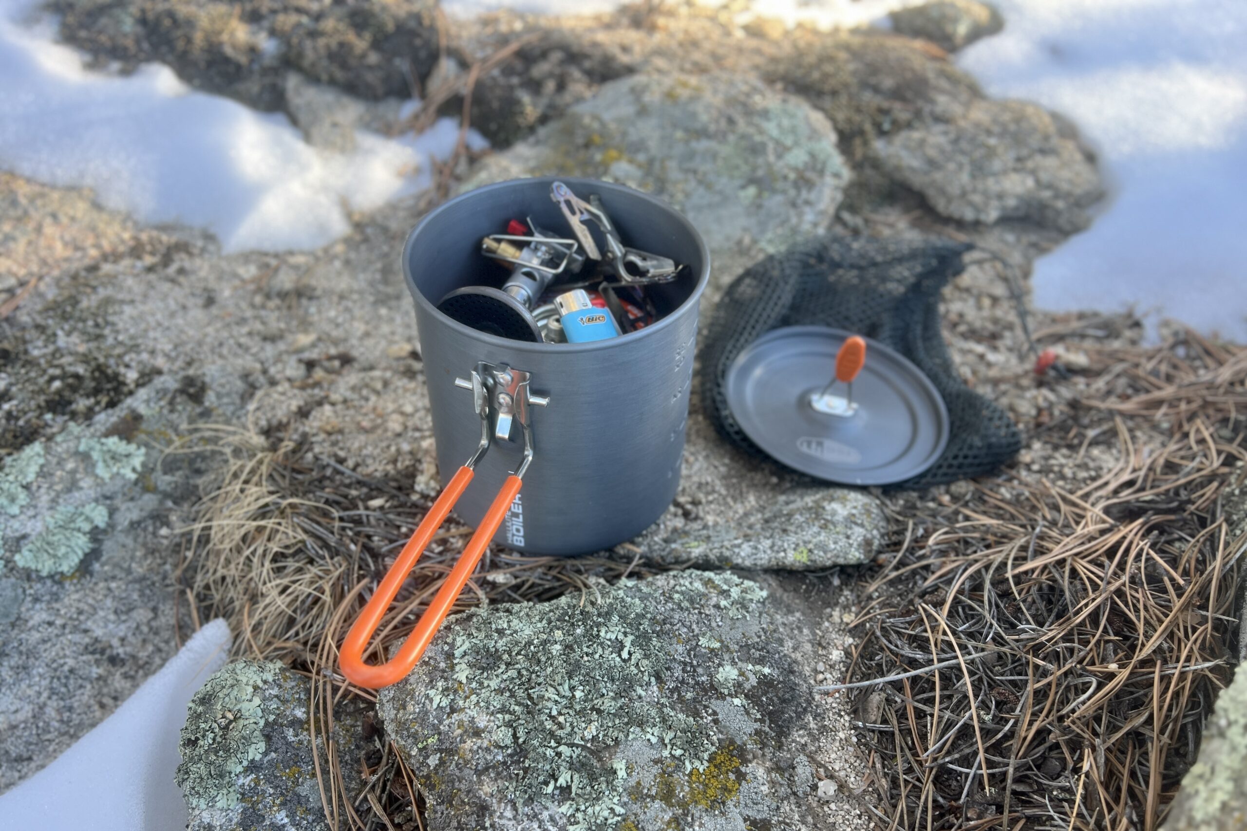 The GSI halulite boiler and lid sitting on a rock with a backpacking stove a lighter packed into the pot.