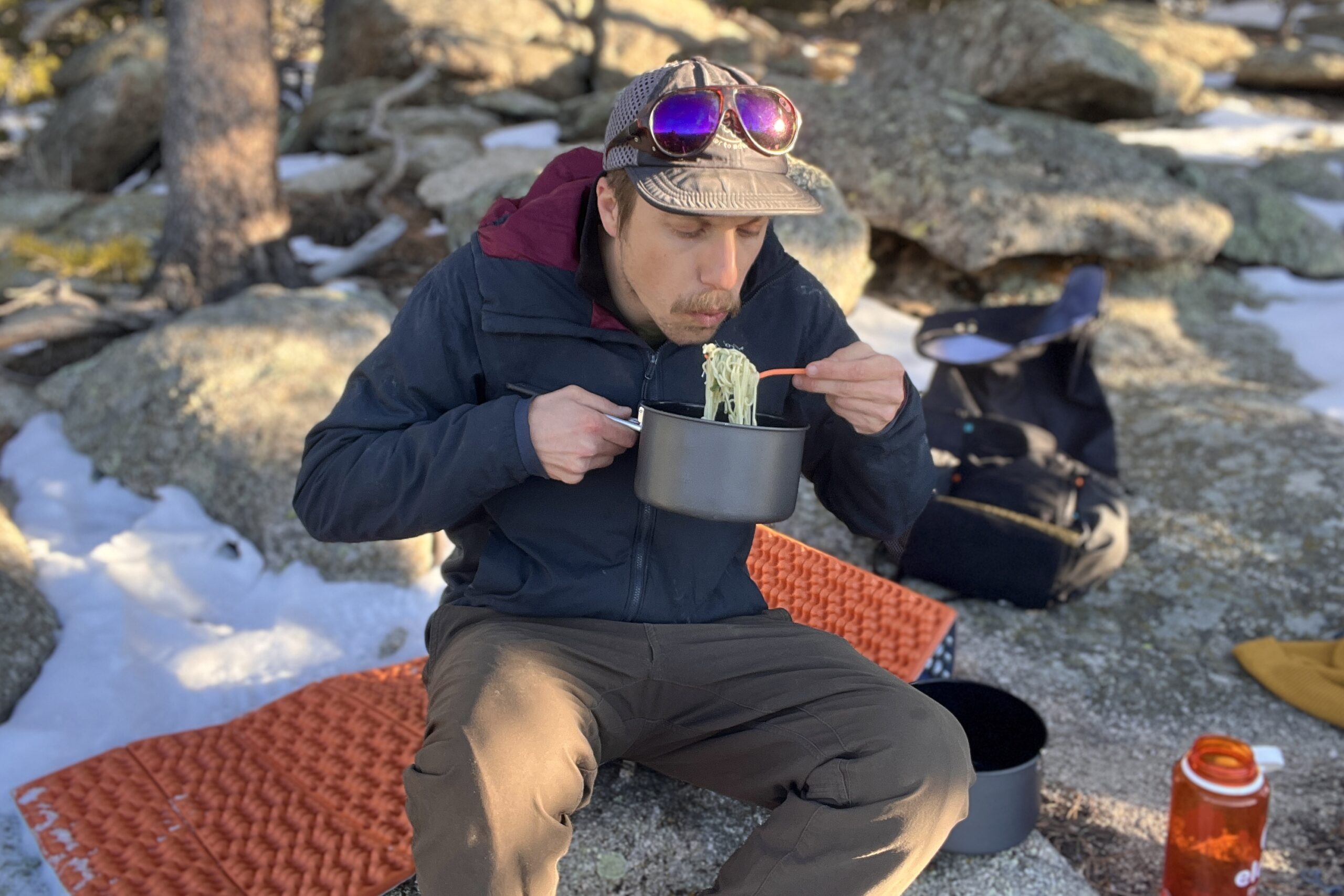 A hiker holds a backpacking pot and blows on a fork full of noodles with rocks and trees in the background.