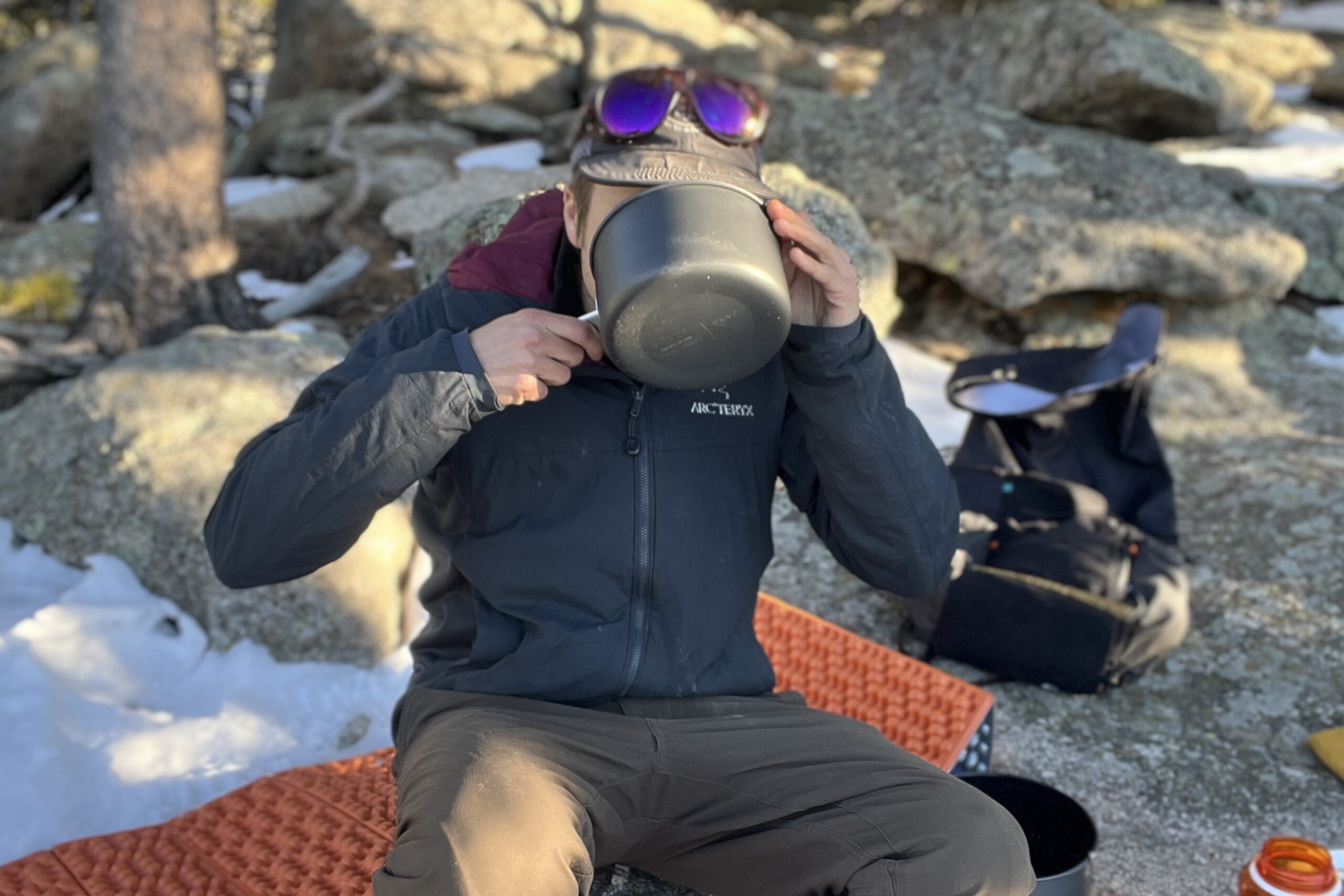 A hiker drinks from a backpacking pot with backpacking gear, rocks, and trees in the background.