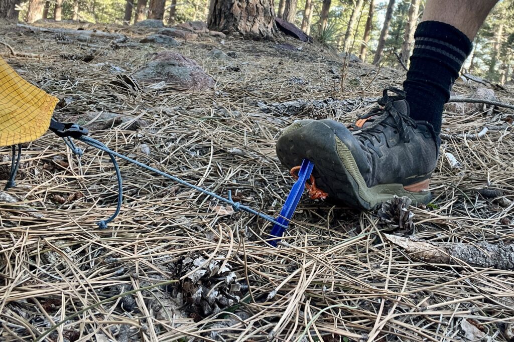 A person steps on a blue tent stake attached to a tent guyline in a forest.