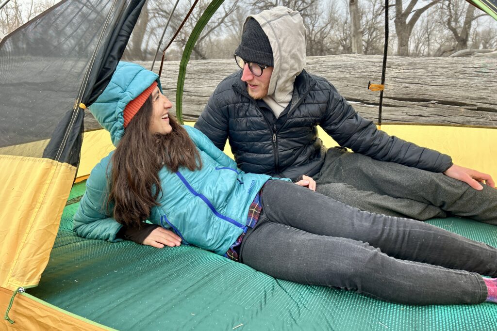 A woman and man are smiling and talking while laying on a camping mattress in a tent in the woods.