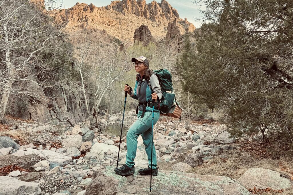 A person is standing on a rock and holding the Distance Carbon trekking poles. The area is rocky with some trees surrounding the wash. There are some rocky, desert peaks in the distance.