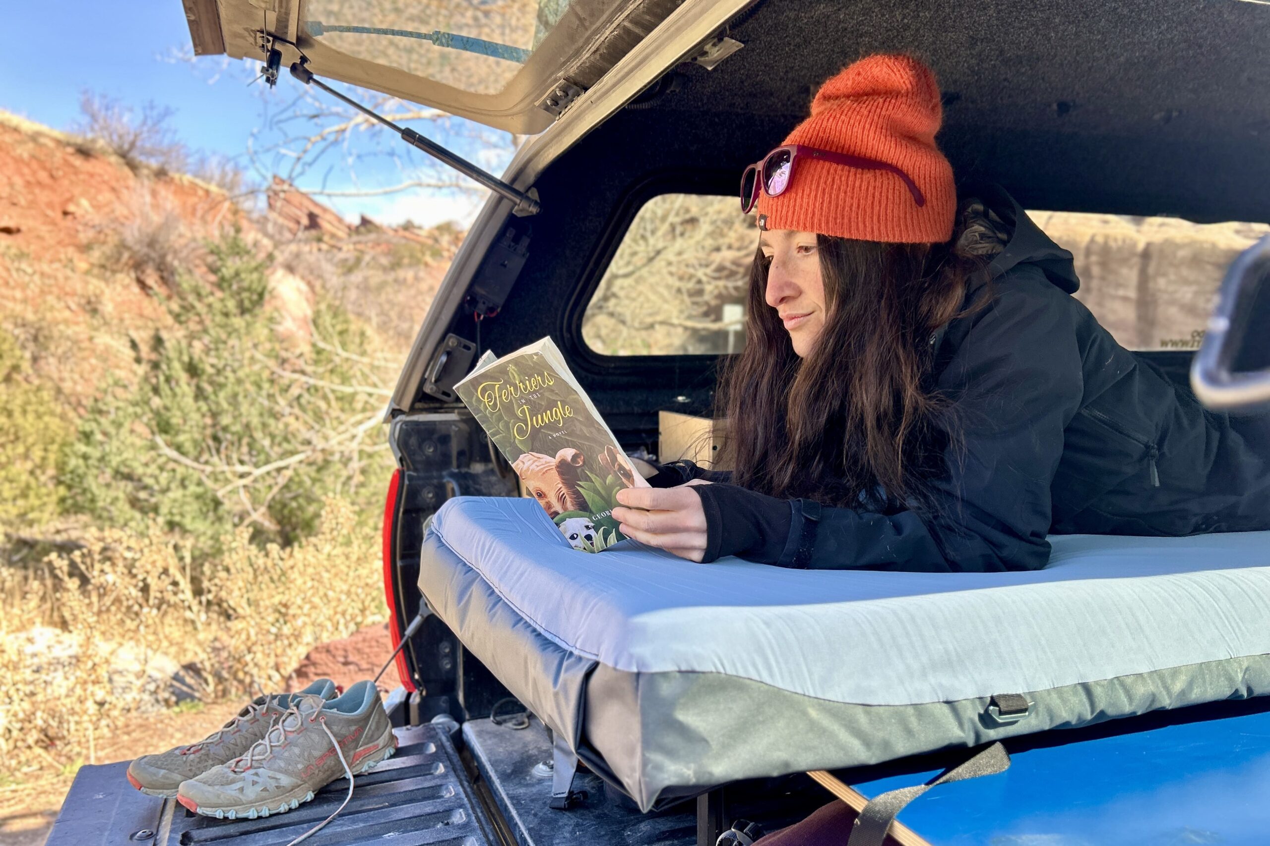 A woman reads a book while laying on a camping mattress in the back of a pickup truck.