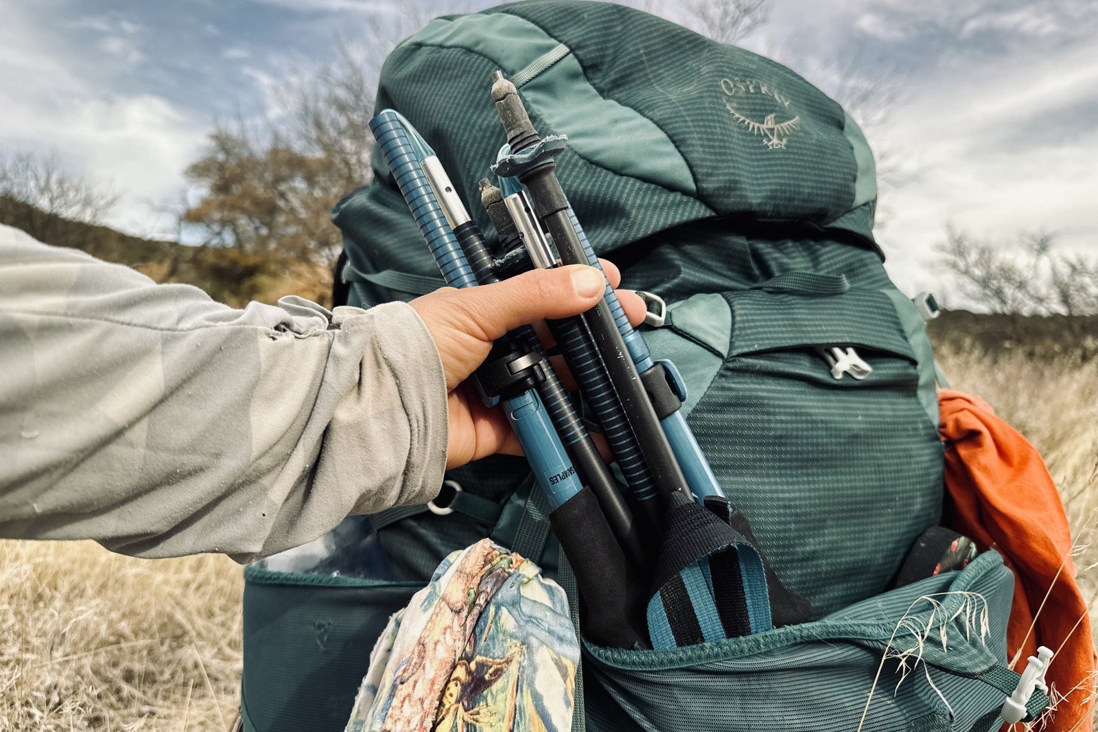 Close up of a hand pulling the folded up Distance Carbon trekking poles from the front pouch of a backpack. There is grass and desert trees in the background.