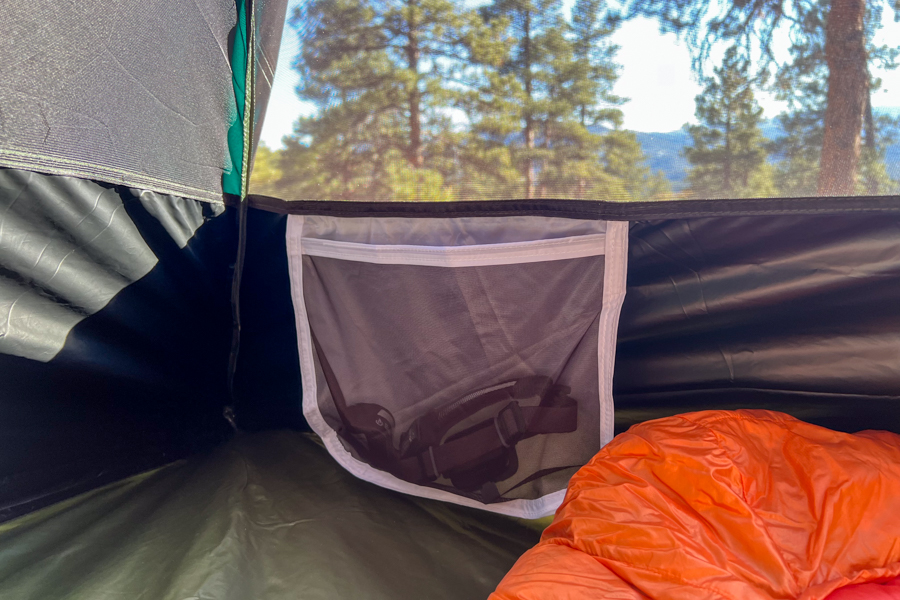 A mesh pocket inside a tent holds a headlamp. You can see pine trees through the mesh body of the tent.