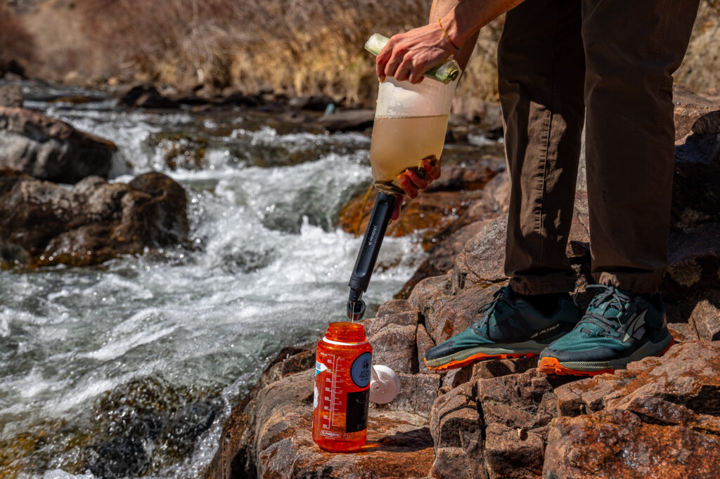 A hiker stands next to a creek and filters dirty water from a bladder, through the peak straw, into a nalgene bottle.