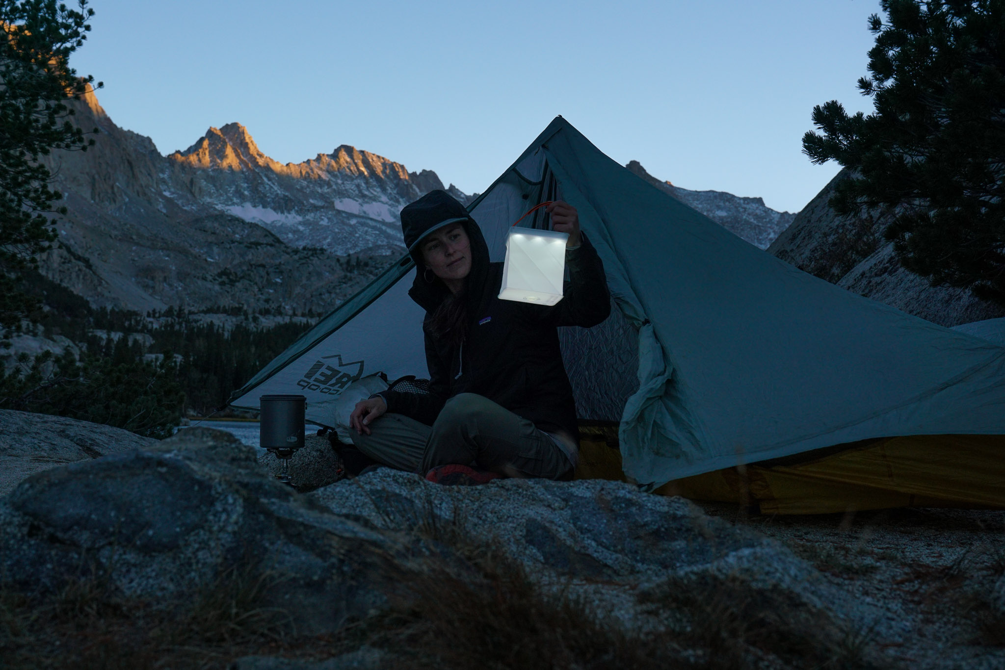 A person holds up a lit LuminAid Packlite Max 2-in-1 lantern. They sit on the ground outside a tent with mountain peaks in the background.