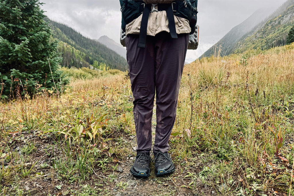 A person is standing in a mountain meadow on a rainy day wearing a soaked pair of the Dynama pants.