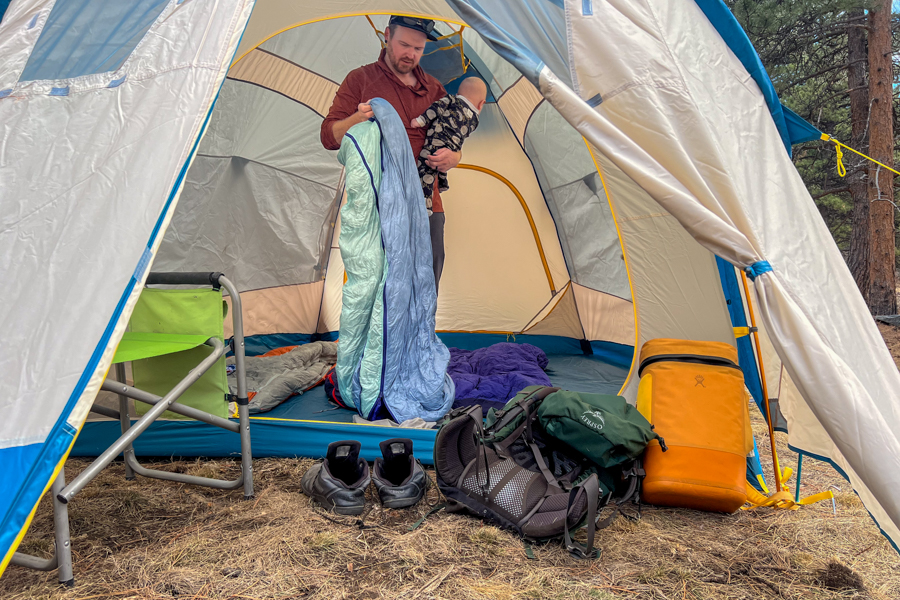 A man and a baby inside a tent. The man is holding a baby and a sleeping bag.