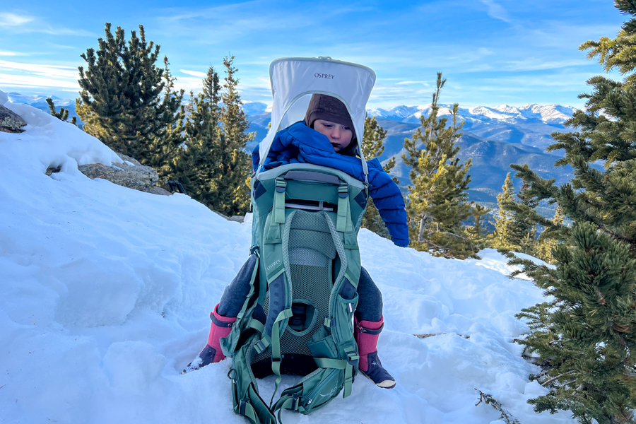A child sitting snugly in the Osprey Poco Premium child carrier on a snowy mountain trail, surrounded by snow-covered trees and distant peaks.