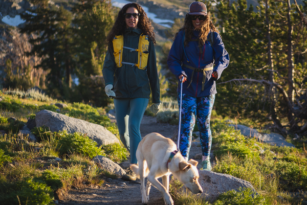 Two women hiking, featuring hiking leggings of two different styles and patterns, one plain blue, and another also blue but camouflage like. Also a small dog, yellow fur. Both women are wearing day packs and rain jackets. The background is green plants and trees. The women are hiking side by side, with the dog in the front.