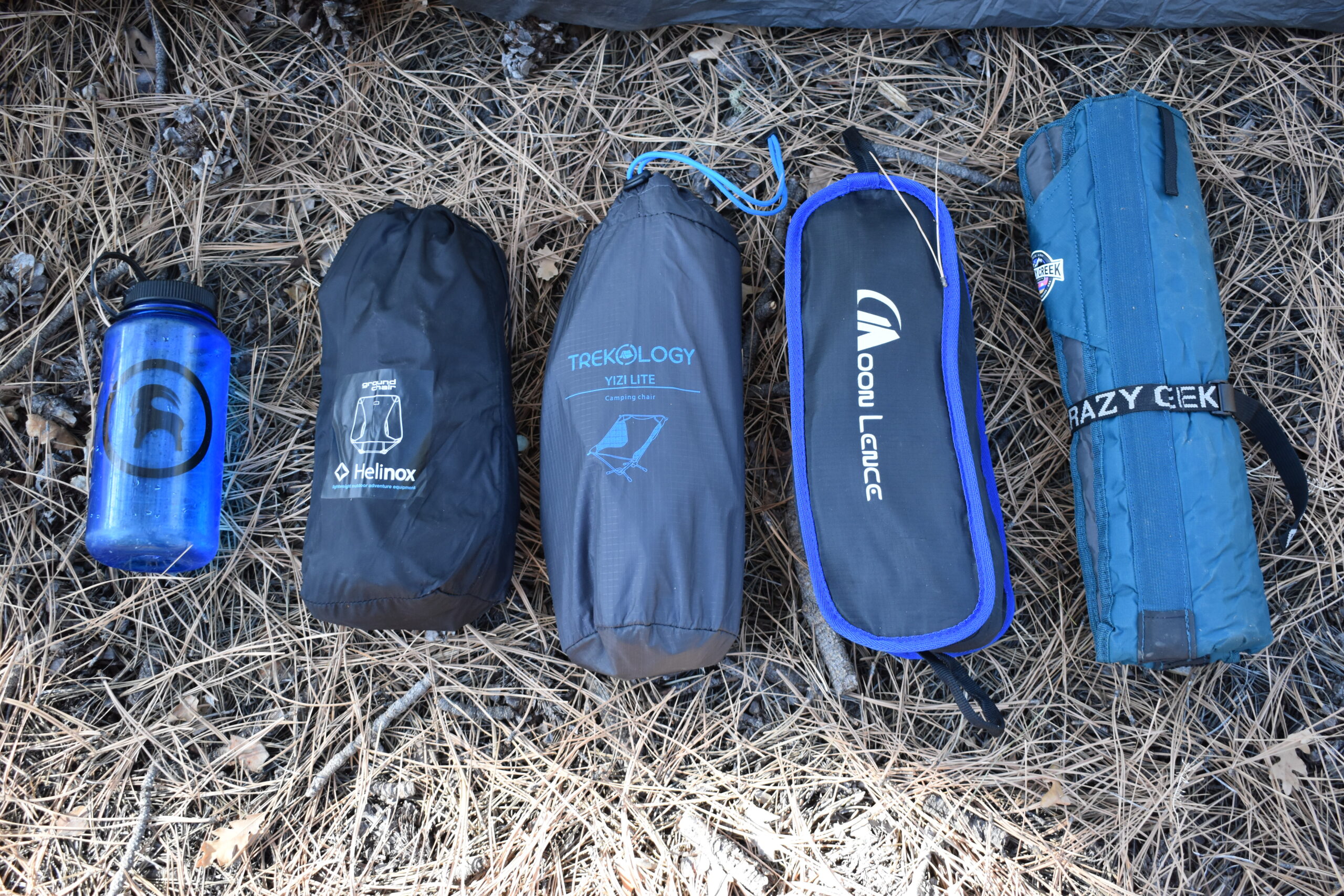 Nalgene water bottle placed next to four backpacking chairs with larger packed sizes: Helinox Ground Chair, Trekology Yizi-Lite, Moon Lence Portable, Crazy Creek Hex 2.0