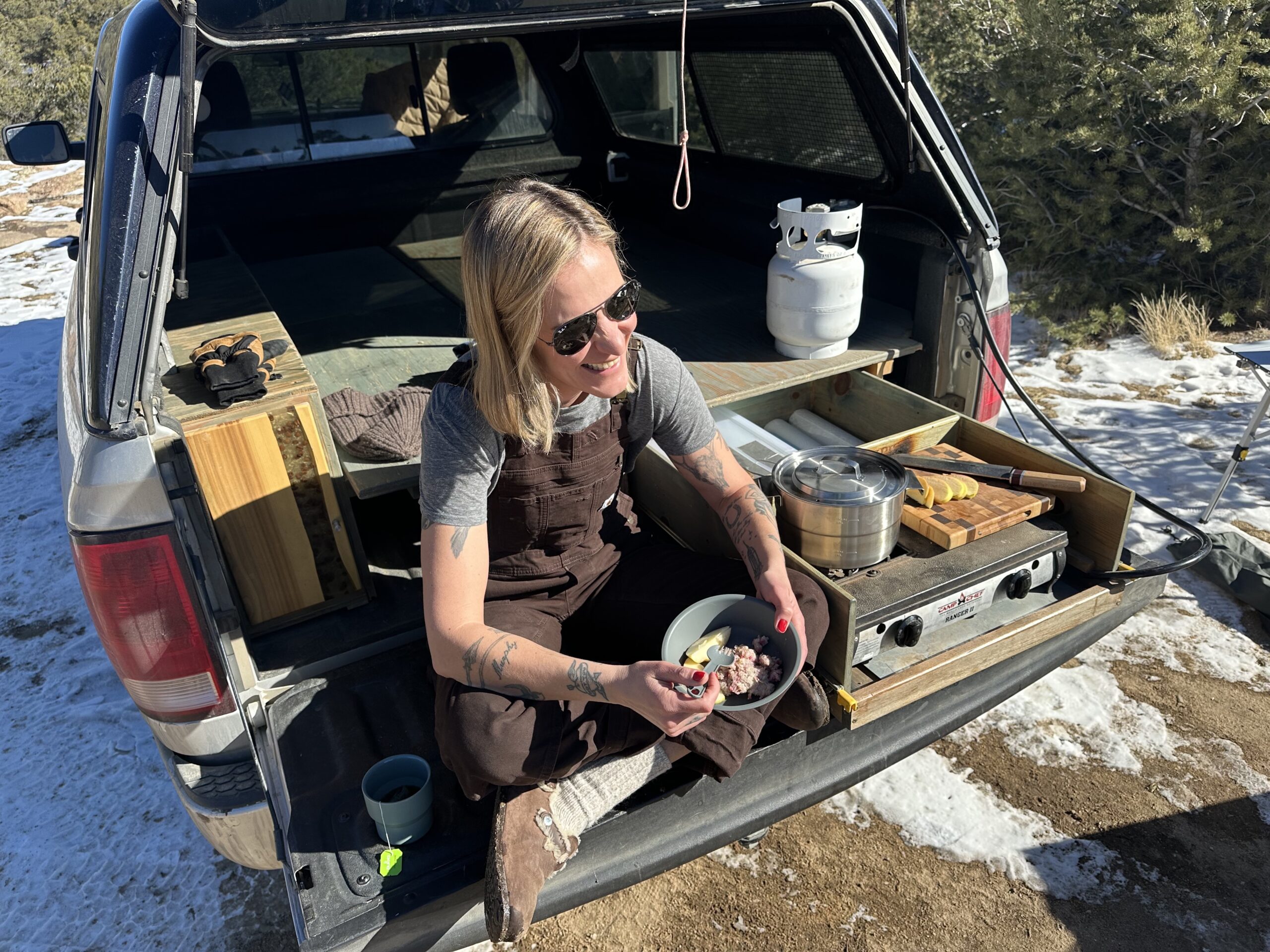 a women wearing overalls sits on the back of her trucks tailgate eating out of the Stanley Wildfare Core Full Serve Cookset
