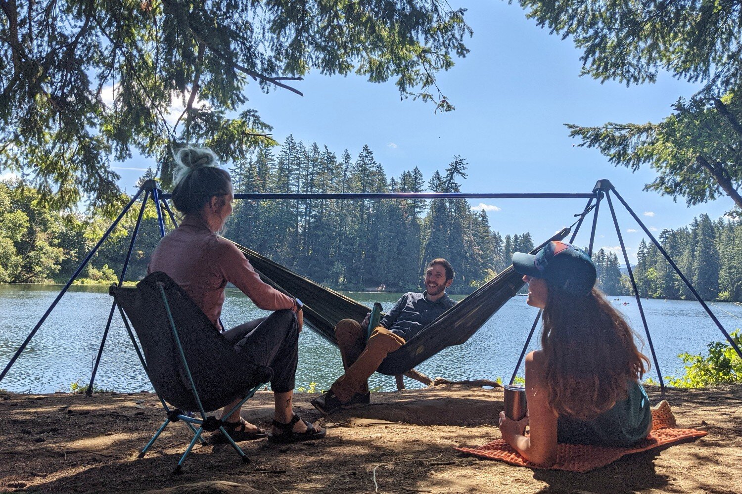 Three people sitting in front of a lake view in a hammock a backpacking chair and sitpad